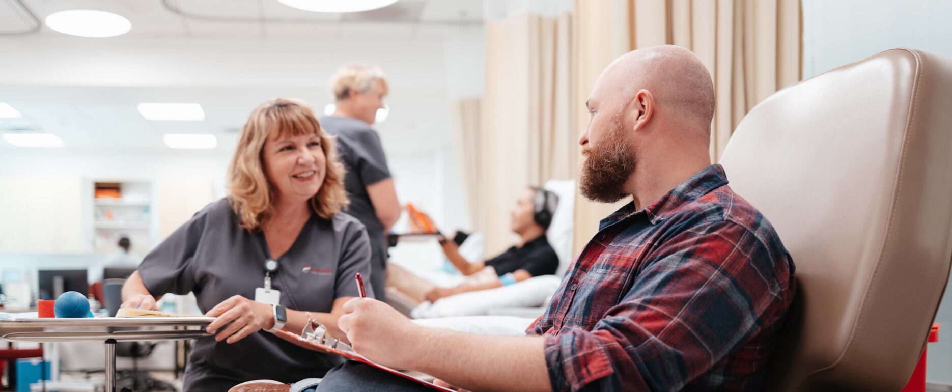 A male patient with a shaved head and short beard sits in a chair being consulted by a nurse with shoulder-length blonde hair in the Canventa office.